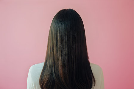 A person stands with their back turned, showing glossy dark long hair. The background features a soft pink color that adds a gentle atmosphere to the indoor space.の素材