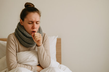 A woman in a neutral sweater and scarf sits on a bed, coughing softly while wrapped in a blanket. The indoor lighting suggests a chilly winter day, creating a cozy atmosphere.の素材
