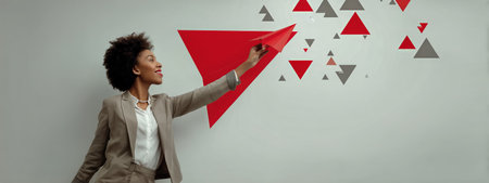 A confident businesswoman in a gray suit playfully throws a red paper airplane towards a wall decorated with colorful triangles in a modern office environment.の素材