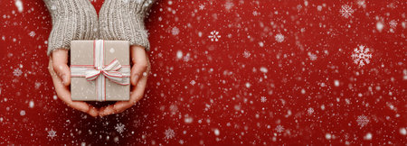 A hand holds a red gift box adorned with a white ribbon while artificial snowflakes gently fall in a warm, festive setting that captures the spirit of the holiday season.の素材