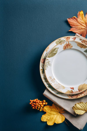 Colorful autumn leaves are arranged on a dark textured surface alongside simple plates and a checkered napkin, creating a warm, seasonal atmosphere for dining.の素材