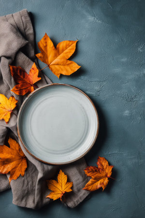 An empty white plate rests on a soft gray cloth surrounded by vibrant autumn leaves, capturing the essence of the season. The blue background adds contrast to the warm colors.の素材