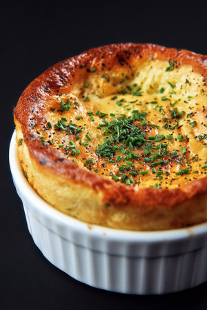 This dish features a fluffy cheese souffl with a golden-brown top, garnished with fresh herbs, served in a white ramekin against a dark background.の素材