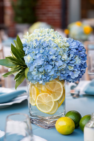 A clear vase filled with blue hydrangeas and lemon slices complements a beautifully set table. Vibrant limes are placed beside the arrangement on a light blue tablecloth.の素材