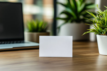 A blank white card sits on a wooden desk, accompanied by a laptop and a small green potted plant, creating a calm and organized atmosphere for work or creativity.の素材
