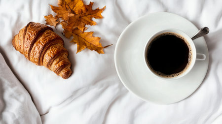 Sipping black coffee from a white cup next to a flaky croissant and fallen autumn leaves on a soft blanket during a calm morning.の素材