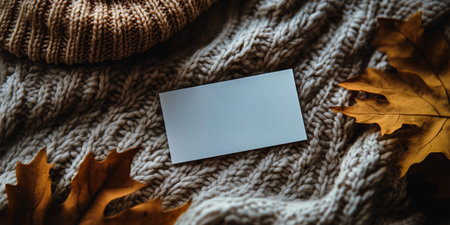 Cozy knitted items including a hat and scarf lie alongside a blank card and dried leaves on a textured surface, creating a warm autumn atmosphere perfect for seasonal notes.の素材