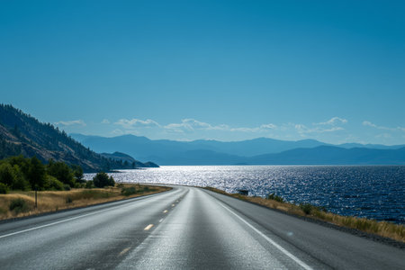 A winding highway runs beside a sparkling lake, surrounded by mountains and green trees, capturing a peaceful afternoon atmosphere under a bright blue sky.の素材