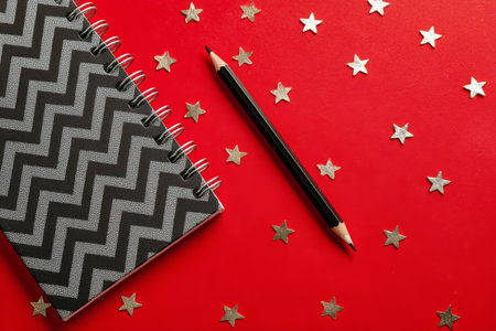 A zigzag patterned notebook rests on a red surface adorned with golden stars. Next to it, a bright red pencil adds a pop of color and creativity to the workspace..の素材