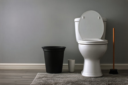 This bathroom features a white toilet, a black waste bin, and a plunger positioned beside a gray rug. The walls are a soft gray, creating a calm atmosphere.の素材