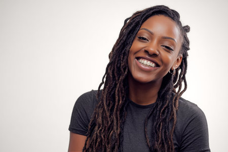A joyful woman with long dreadlocks smiles, showing her natural beauty. She wears a simple black top and stands against a neutral background, radiating happiness and confidence..の素材