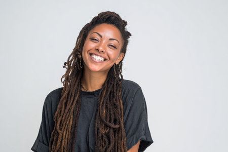 A joyful woman with long dreadlocks smiles, showing her natural beauty. She wears a simple black top and stands against a neutral background, radiating happiness and confidence.の素材