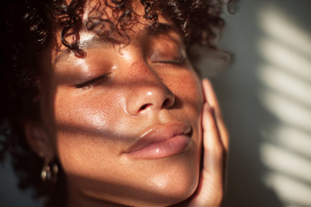 A woman with natural curls rests her head against her hand, eyes closed in peaceful solitude. Soft sunlight filters through blinds, creating a warm and calming atmosphere.の素材