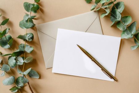 A blank card and envelope are placed on a soft beige surface. A gold pen lies nearby, while fresh eucalyptus leaves add a natural touch to the scene.の素材