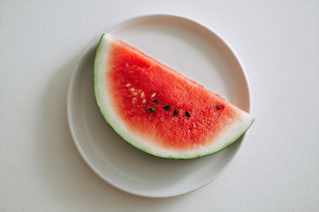 A refreshing slice of juicy watermelon rests on a plain white plate, showing its vibrant pink interior and green rind. The bright lighting enhances the fruits colors.の素材