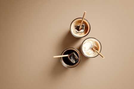 Three stylish glasses of iced coffee are arranged on a beige surface, each with different layers of milk and coffee. Wooden straws add a rustic touch to the refreshing drinks.の素材