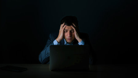 A man sits at a desk with a laptop, looking stressed and deep in thought. The room is dark except for a lamp casting light on his focused expression and surroundings.の素材