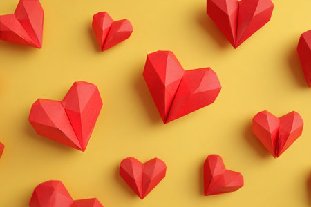 Colorful red paper hearts of various sizes are beautifully scattered on a cheerful yellow background, ideal for festive decoration.の素材