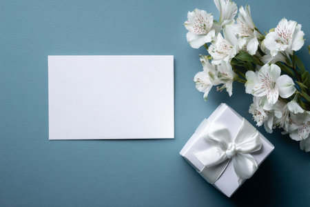 A small, neatly wrapped gift sits next to a bunch of white flowers and a blank card on a soft blue surface, perfect for celebrations, thank you notes, or personal messages.の素材