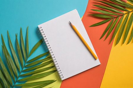 A blank notebook lays on a vibrant, multicolored background with a pencil beside it. Fresh green leaves add a touch of nature, inviting creativity and inspiration.の素材