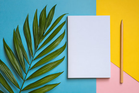 A blank notebook lays on a vibrant, multicolored background with a pencil beside it. Fresh green leaves add a touch of nature, inviting creativity and inspiration. AI Generativeの素材