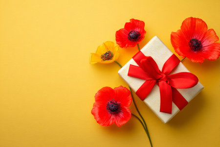A neatly wrapped gift box with a red ribbon is placed beside bright orange and red poppy flowers against a cheerful yellow backdrop, creating a festive atmosphere. AI Generativeの素材