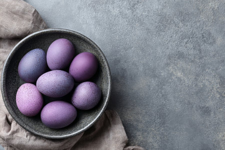 A bowl holds several dyed purple eggs arranged neatly. The bowl is placed on a soft cloth on a smooth gray surface, perfect for spring celebrations.の素材