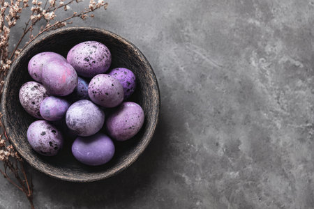 A wooden bowl holds various speckled eggs in shades of purple and gray, arranged neatly on a textured dark surface. The scene suggests a festive or springtime theme. AI Generativeの素材