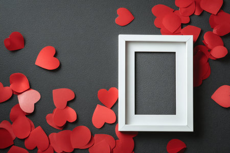 A white picture frame sits empty on a dark surface, surrounded by scattered red paper hearts. This setup creates a romantic and artistic atmosphere ideal for celebrations.の素材