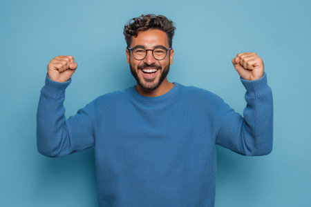 A man with curly hair and glasses is smiling widely, raising his arms in celebration. He is wearing a cozy blue sweater and stands in front of a solid blue backdrop.の素材
