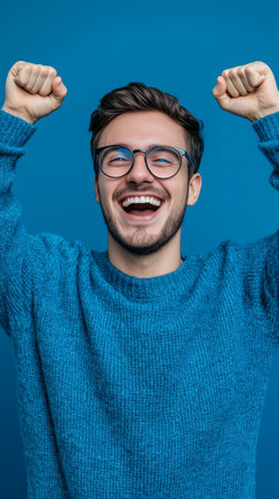 A young man with glasses is smiling widely, raising his arms in excitement against a bright blue backdrop. He wears a knit sweater and looks very happy.の素材