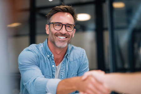A confident man wearing glasses smiles as he shakes hands with a colleague in a contemporary office environment during a business meeting.の素材