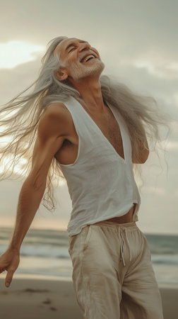 A man with long silver hair is standing on a beach, smiling and embracing the moment as the sun sets. The waves are gently crashing in the background.の素材
