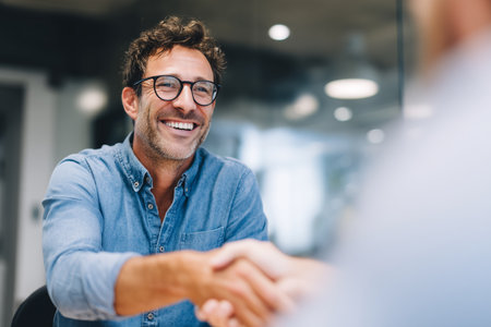 Two men engaged in a business meeting inside a modern office. One man is smiling while shaking hands, conveying a sense of agreement and positivity in their discussion.の素材