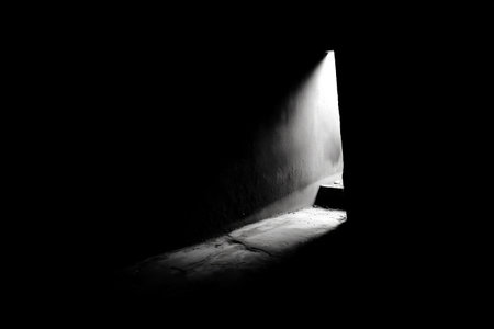 A narrow beam of light shines from an open door, illuminating the dark hallway. The contrast highlights the textures on the walls and floor, creating a moody atmosphere.の素材