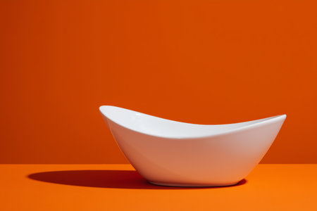 A sleek white bowl is placed against a vibrant orange backdrop, highlighting its smooth curves and minimalistic style. The scene emphasizes modern design and color contrast.の素材