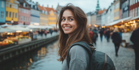 In a vibrant city, a young woman with long hair stands by the water, smiling brightly. Colorful buildings surround the area, creating a lively atmosphere. People stroll nearby.の素材