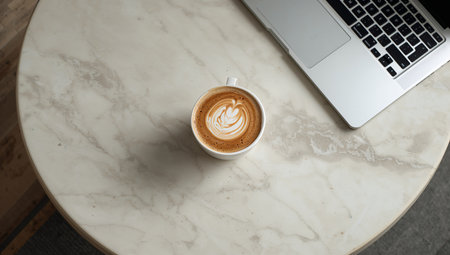 A white coffee cup with latte art sits on a marble table next to a laptop. The scene creates a warm and inviting workspace atmosphere perfect for productivity.の素材
