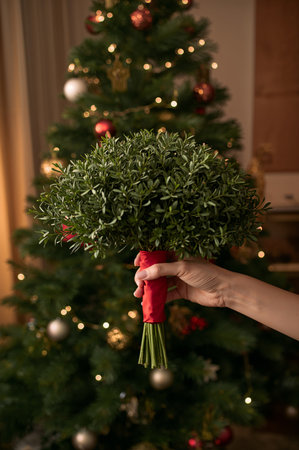 A hand presents a lush greenery bouquet tied with a red ribbon, standing near a decorated Christmas tree adorned with golden ornaments and twinkling lights.の素材
