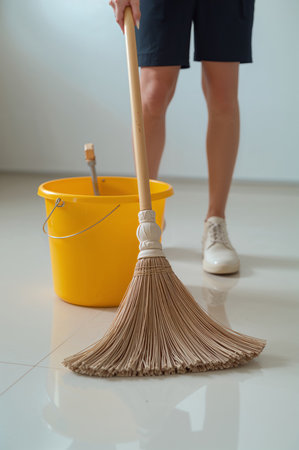 Person sweeps the floor with a broom while standing next to a yellow bucket. The clean, spacious room is filled with natural light during the afternoon.の素材