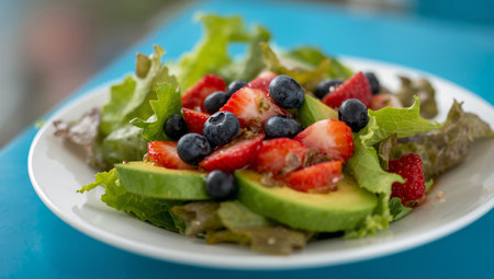 Fresh salad made with greens, strawberries, blueberries, and avocado sits on a white plate on a blue wooden surface. The vibrant colors create an inviting presentation.の素材