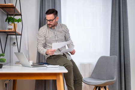 Attractive young Caucasian manager sitting on the desk reviewing latest sales report while working...
