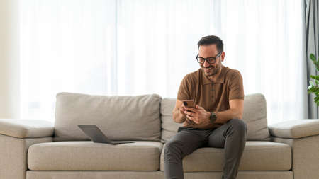 Excited adult man using phone for online payment shopping online from home, Casual entrepreneur working at home, sending text message to his coworkerの写真素材