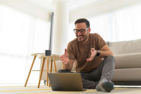 Happy adult man working at home sitting on the floor talking at online meeting with his coworkers about important project satisfied with their solution for problemの写真素材