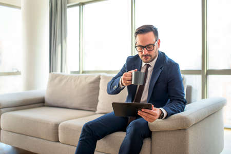 Confident young businessman sitting at sofa having a coffee break reading email on tablet computer. Concept of successful young entrepreneur enjoying his moments of silence drinking coffeeの写真素材