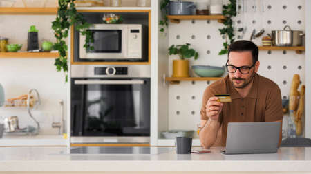 younger man preparing to make a reservation for vacation leaning on the counter in the kitchen using...