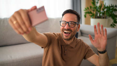 Handsome middle aged man using smart phone video call waving hand looking at camera and talking with his friendsの写真素材