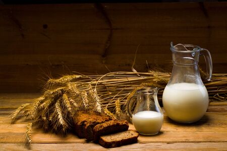 Still life with a glass jug of milk, a glass mug, rye bread, ears of corn on the background of wooden boardsの写真素材