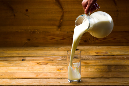 Milk from a glass jug poured into a glass, a loaf of rye bread, ears of corn on the background of wooden boards, studio lightの写真素材