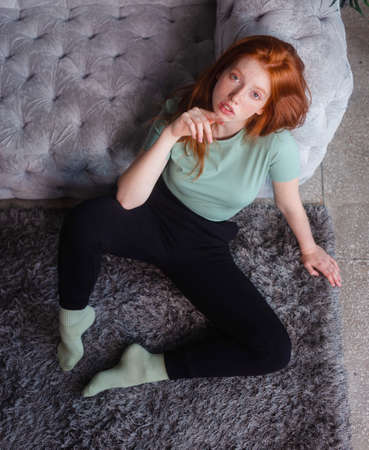Portrait of a beautiful redhead girl sitting in casual clothes on floor near couch spending time in...